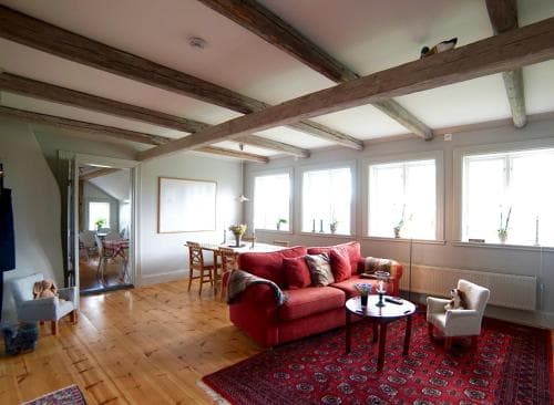A cozy living room with exposed wooden beams, a vibrant red sofa, and large windows inviting natural light, ideal for travelers seeking a warm and inviting accommodation experience.