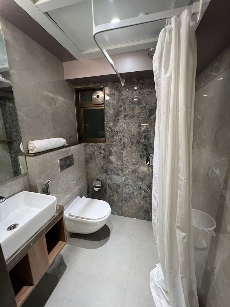 A modern hotel bathroom in Mumbai features sleek marble walls, a white toilet, a sink with wooden accents, and a shower area with a white curtain, ideal for travelers seeking comfort and cleanliness.