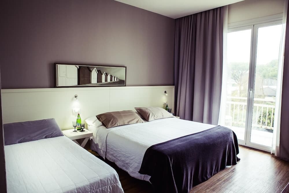 A cozy hotel room with purple accents, two neatly made beds, a wall-mounted mirror, and a balcony offering a serene outdoor view, ideal for travelers seeking comfort and relaxation.