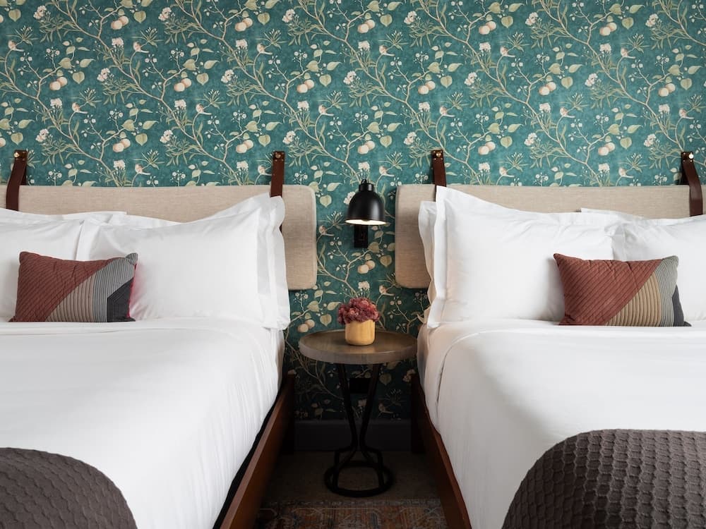 A cozy hotel room features two neatly made beds with white linens and patterned pillows against a vibrant floral wallpaper backdrop, complemented by a small wooden table with a flower vase, ideal for travelers seeking comfort and charm.
