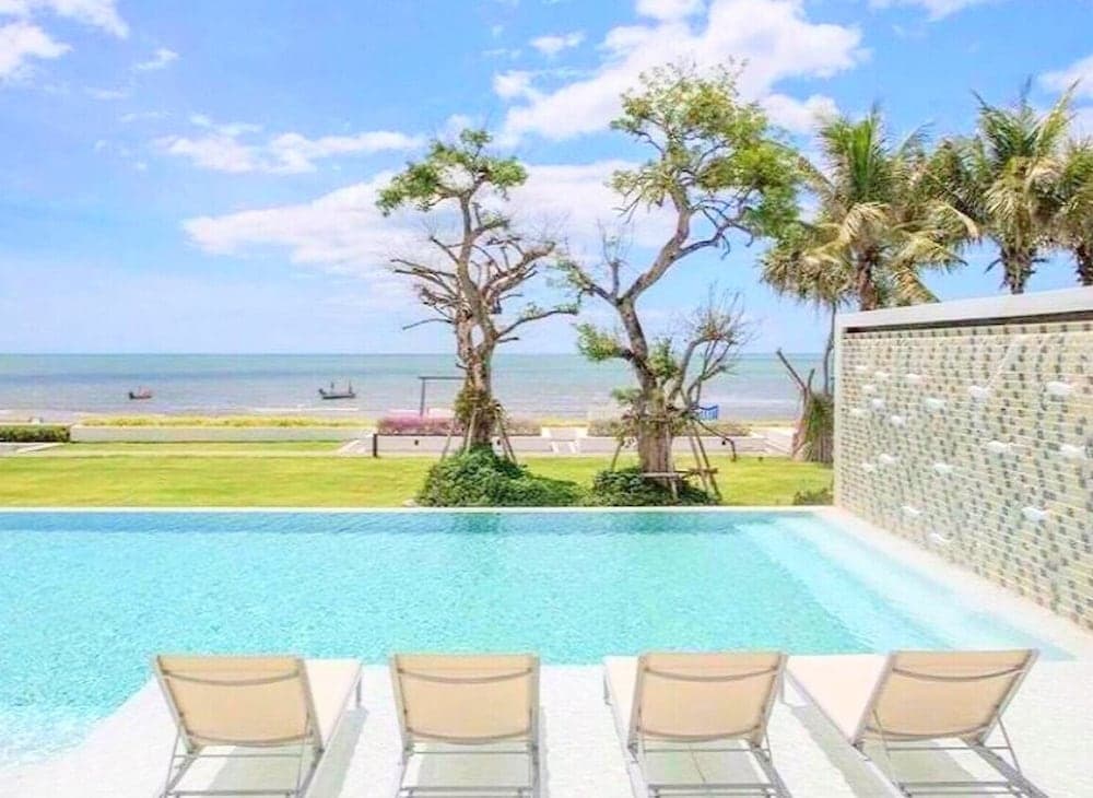 A serene beachside resort features a tranquil infinity pool lined with lounge chairs, lush greenery, and palm trees under a bright blue sky, ideal for luxury relaxation and vacation photography.