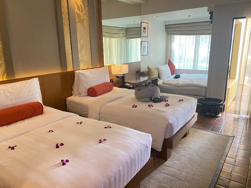 A cozy hotel room at SAii Laguna Phuket features neatly arranged twin beds adorned with purple flower petals, a serene ambiance, and large windows inviting natural light, perfect for travelers seeking relaxation in Thailand.