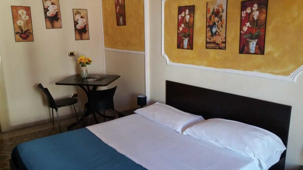 A cozy hotel room features a neatly made bed, a small dining table with chairs, vibrant floral artwork on the walls, and a warm ambiance ideal for travelers seeking comfort and relaxation.