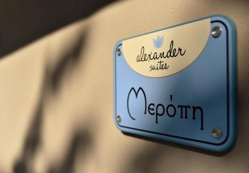A close-up of a stylish blue sign reading "Alexander Suites" with Greek script below, set against a textured wall, evokes a modern and elegant hotel ambiance ideal for travelers seeking upscale accommodations.