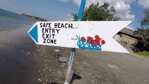 A vibrant signpost marking the "Safe Beach Entry Exit Zone" stands on a sandy shore by calm waters, ideal for travelers seeking secure beach access amidst a serene coastal setting.