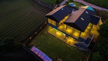 A cozy, warmly lit house nestled amidst lush green fields at dusk offers a serene retreat, ideal for travelers seeking relaxation in a tranquil countryside setting.