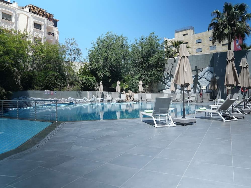 A serene urban poolside scene features lounge chairs, closed umbrellas, and lush greenery against a backdrop of modern buildings, ideal for travelers seeking relaxation in a city retreat.