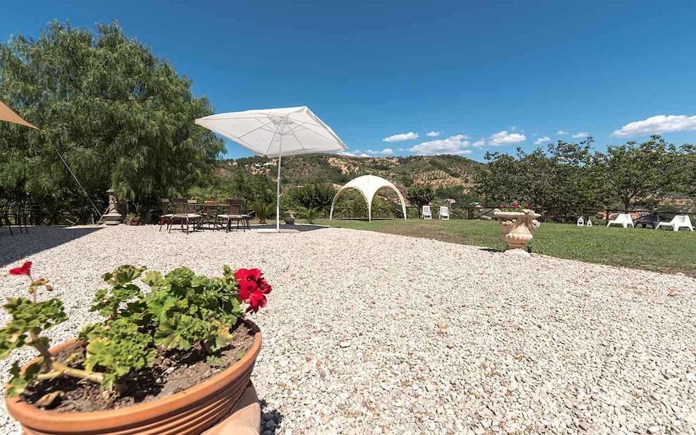 A serene outdoor retreat features a gravel patio with potted red flowers, a white umbrella over seating, and a scenic backdrop of rolling hills under a clear blue sky, ideal for relaxing vacations or leisurely getaways.