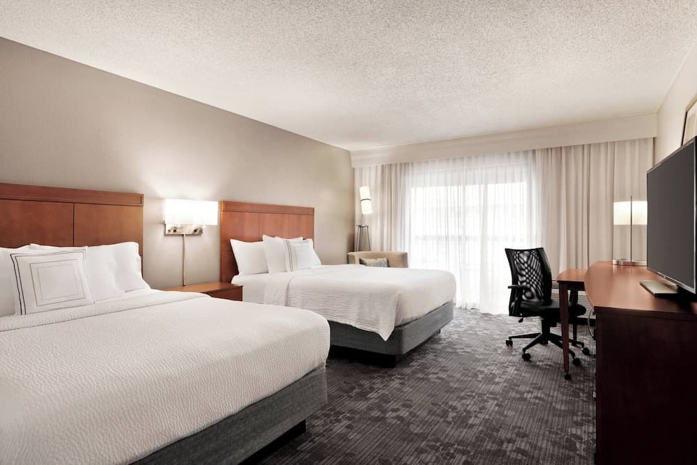A cozy hotel room features two neatly made beds, a desk with a chair, and large windows with sheer curtains, creating a welcoming atmosphere ideal for business travelers seeking comfort and functionality.