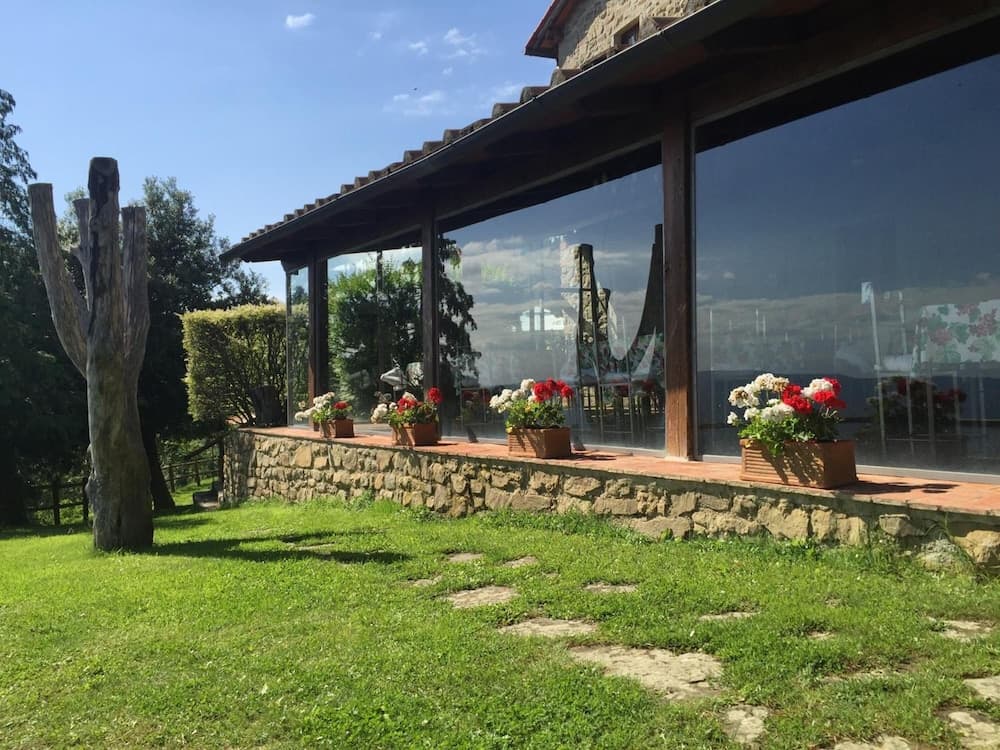 A serene countryside villa features a stone wall lined with vibrant flower pots under a glass-enclosed porch, surrounded by lush greenery, ideal for tranquil retreats and nature-inspired getaways.