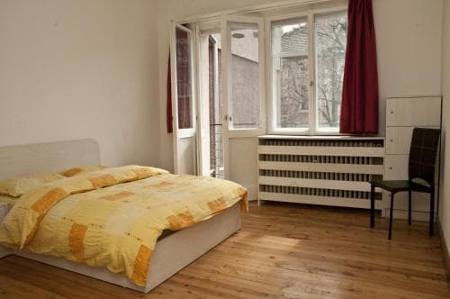 A cozy bedroom with wooden floors, a yellow bedspread, and a view of urban buildings through large windows, ideal for travelers seeking a simple and affordable accommodation option.