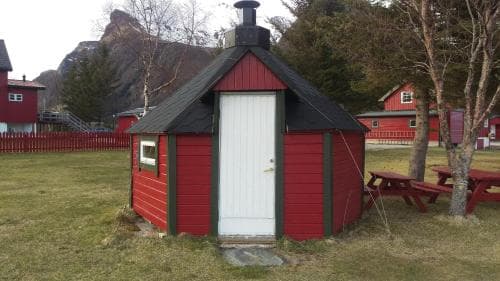 A quaint red and black cabin with a white door sits in a serene rural setting near picnic tables, ideal for travelers seeking a cozy retreat in a peaceful countryside environment.