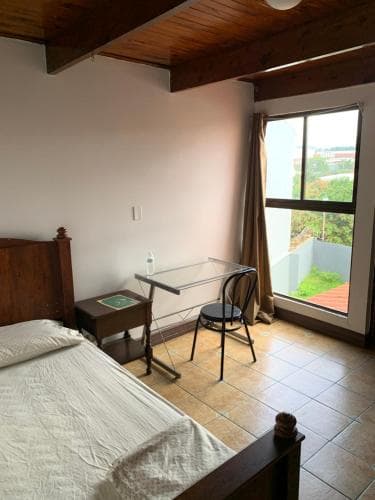 A cozy bedroom with wooden beams, tiled floors, and a large window overlooking greenery offers a serene retreat for travelers seeking simplicity and comfort.