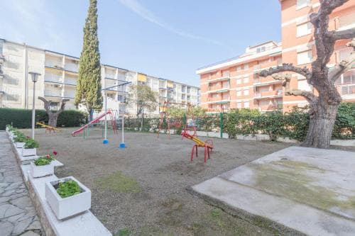 A sunny playground surrounded by residential buildings features colorful equipment, greenery, and a tranquil atmosphere, ideal for family-friendly urban getaways.
