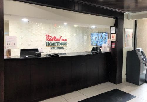 A modern hotel lobby featuring the Red Roof Inn Hometowne Studios reception desk with a sleek black counter, digital displays, and an ATM, ideal for travelers seeking a welcoming and efficient check-in experience.
