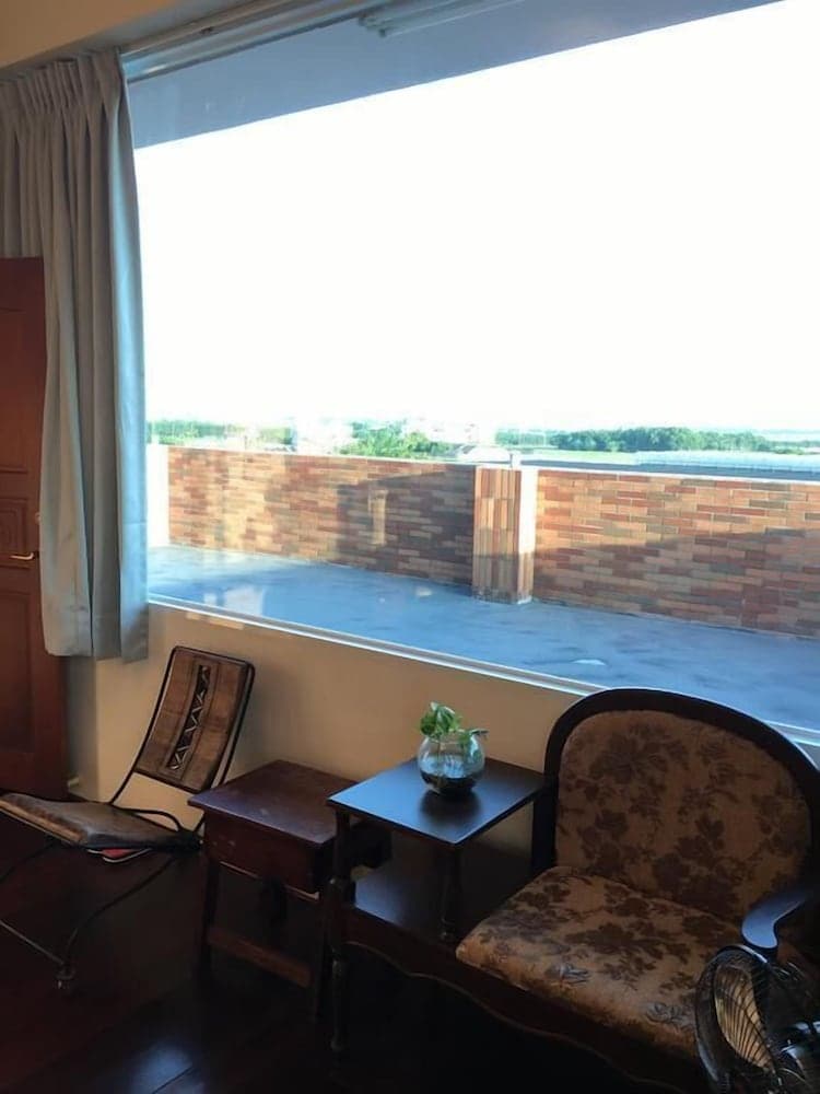 A cozy hotel room with vintage furniture and a small plant on a table offers a serene view of a brick wall and greenery through a large window, ideal for travelers seeking relaxation and comfort.