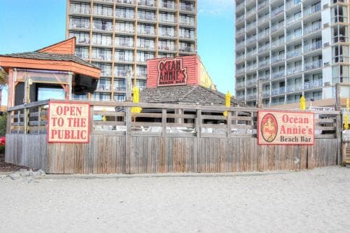 A sandy beachfront scene features Ocean Annie's Beach Bar, a wooden structure with vibrant signage, set against towering condominiums, inviting travelers to enjoy a casual seaside escape.
