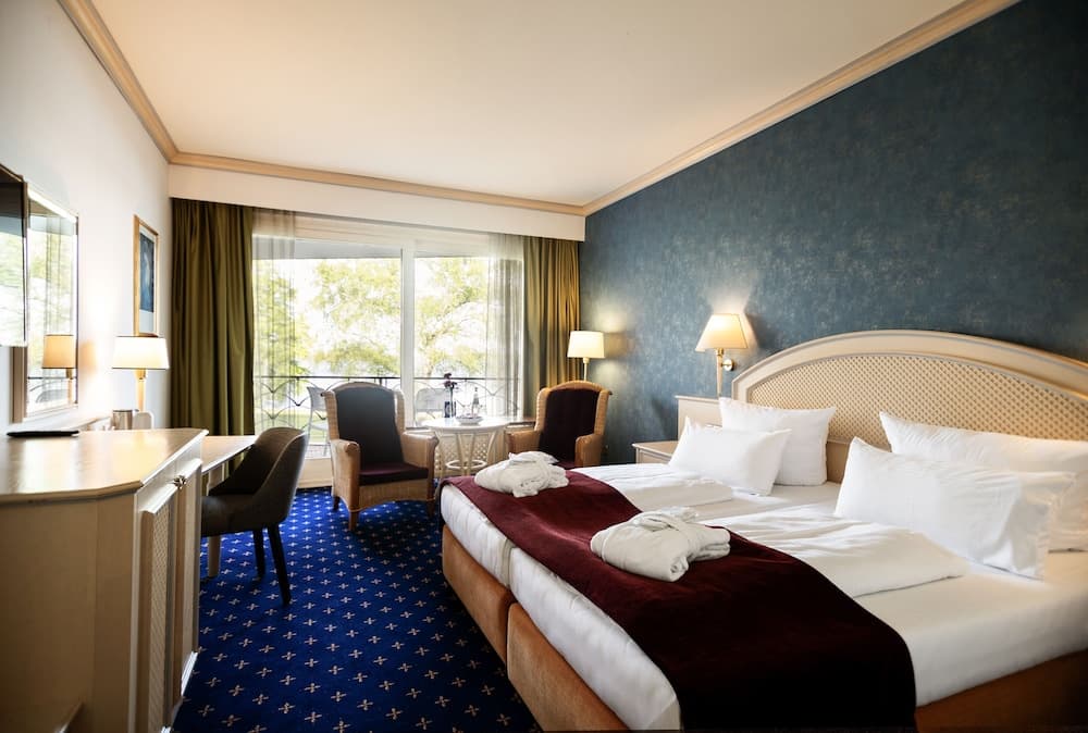 A cozy hotel room features a neatly made bed with plush pillows, a vibrant blue carpet, a desk area by the window, and a balcony view of greenery, ideal for travelers seeking comfort and relaxation.