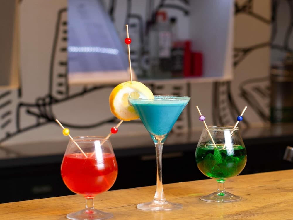 A vibrant trio of colorful cocktails garnished with skewers sits invitingly on a wooden bar counter against an artistic mural backdrop, perfect for a lively night out or cocktail enthusiasts exploring unique drink spots.