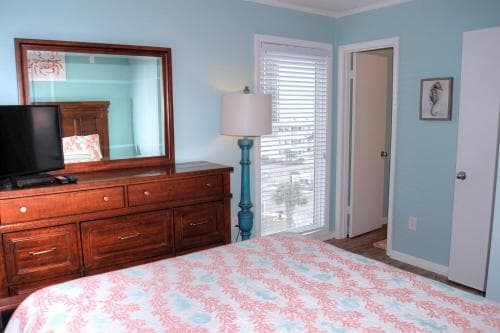 A cozy bedroom with light blue walls, a wooden dresser topped with a TV, a floral bedspread, and a window with blinds offering a serene view, ideal for travelers seeking a relaxing retreat.