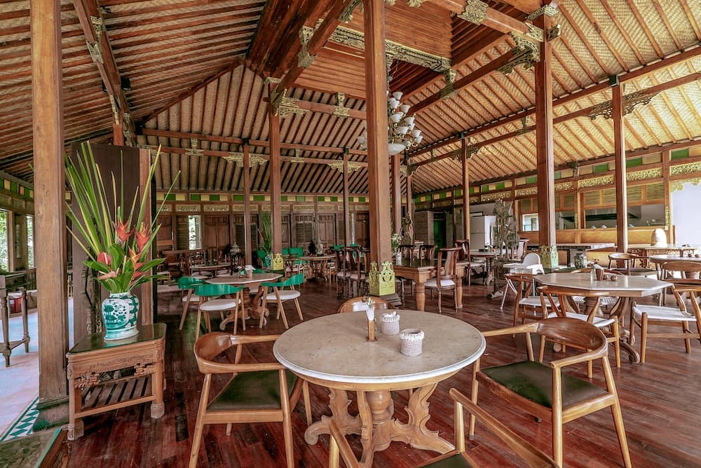 A serene tropical restaurant with wooden beams, thatched roof, and vibrant greenery offers a tranquil dining experience ideal for travelers seeking cultural immersion.