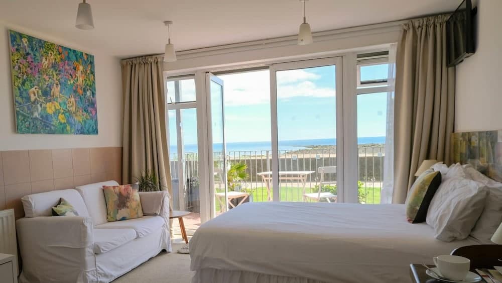 A cozy beachside bedroom with a serene ocean view through large glass doors, featuring comfortable furnishings, vibrant artwork, and a tranquil atmosphere ideal for a relaxing getaway.