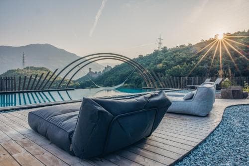 A serene sunset illuminates a luxurious outdoor lounge area beside a pool, featuring plush bean bags on a wooden deck surrounded by lush green hills, ideal for a relaxing getaway or vacation retreat.