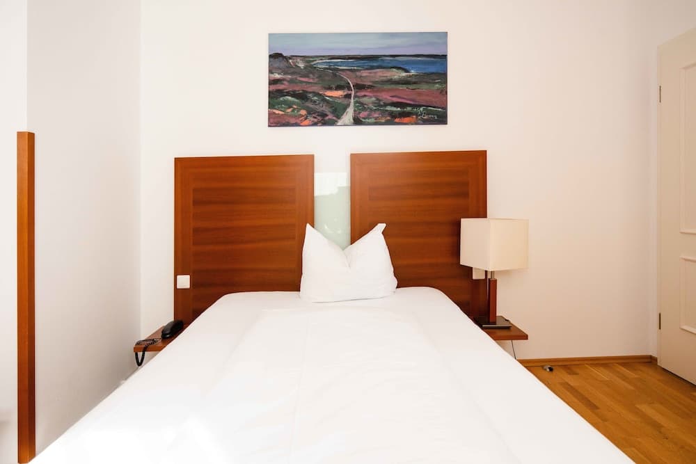 A serene hotel room features a neatly made bed with white linens, wooden headboards, a modern lamp, and an abstract landscape painting, ideal for travelers seeking comfort and simplicity.