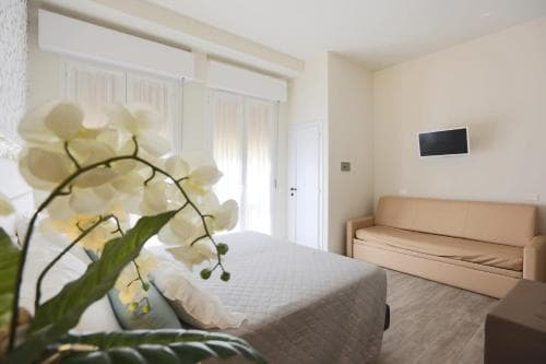 A serene hotel room features a cozy bed adorned with white orchids, a comfortable sofa, and soft natural light filtering through sheer curtains, ideal for travelers seeking relaxation and comfort.