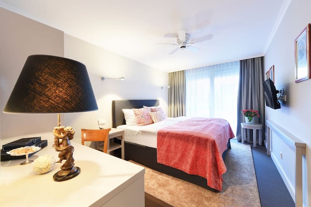 A cozy hotel room features a neatly made bed with a red throw, a stylish desk adorned with a decorative lamp and sculpture, large windows with sheer curtains, and a calming ambiance ideal for travelers seeking comfort and relaxation.