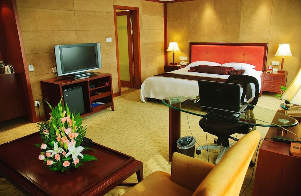 A cozy hotel room features a neatly made bed, a desk with a laptop, a TV on a wooden stand, and a vibrant floral arrangement on the coffee table, ideal for travelers seeking comfort and functionality.