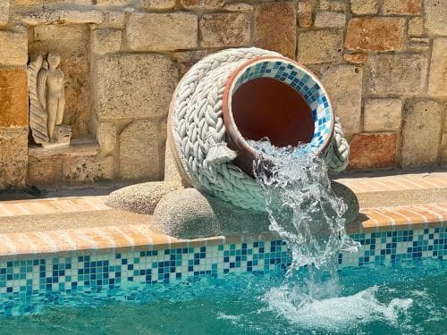 A serene poolside scene features a large ceramic jug pouring water into a mosaic-tiled pool, set against a rustic stone wall with a classical statue niche, ideal for a relaxing vacation retreat.
