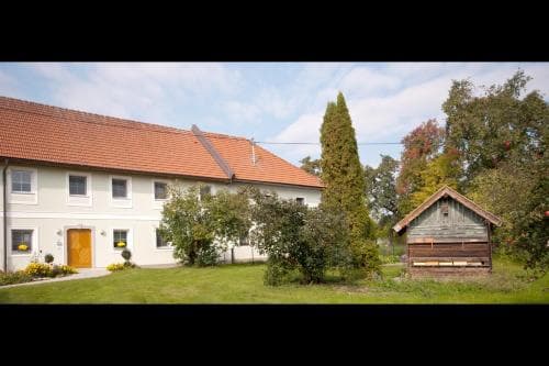 A serene countryside retreat features a charming white farmhouse with a red-tiled roof surrounded by lush greenery, a quaint wooden shed, and vibrant autumn trees, ideal for a peaceful rural getaway.