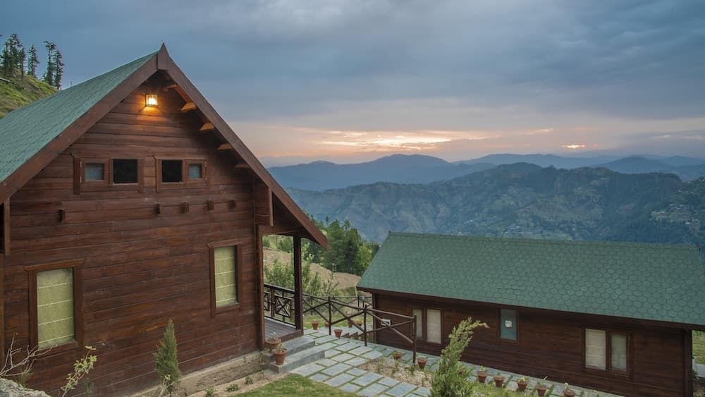 A cozy wooden cabin with a green roof overlooks a serene mountain landscape at dusk, ideal for travelers seeking a tranquil retreat in nature.