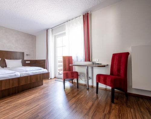 A cozy hotel room with wooden flooring, a neatly made bed, red velvet chairs, and a desk by the window, ideal for travelers seeking comfort and functionality during their stay.