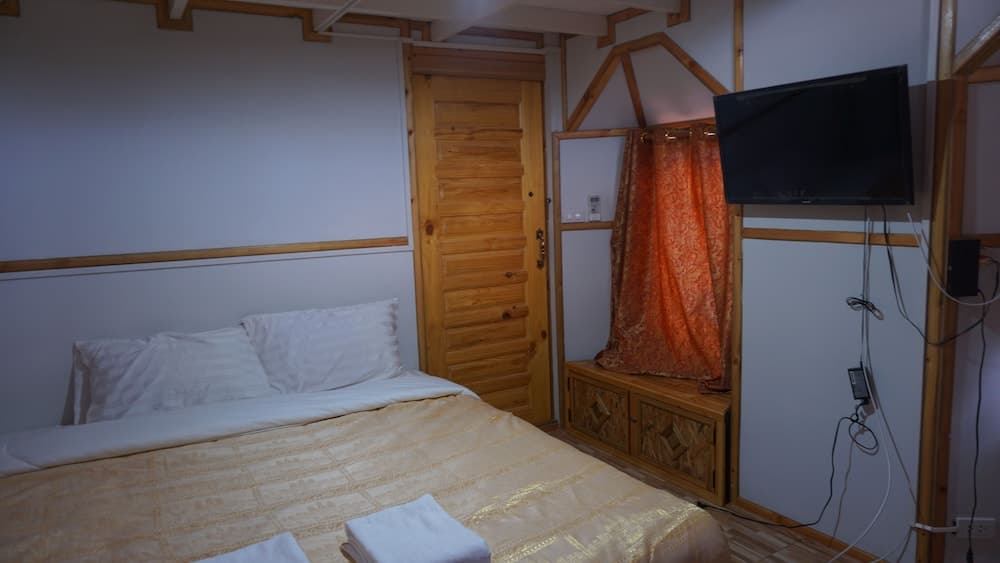 A cozy hotel room features a neatly made bed, wooden accents, an orange curtain, and a mounted TV, ideal for travelers seeking comfort and simplicity.