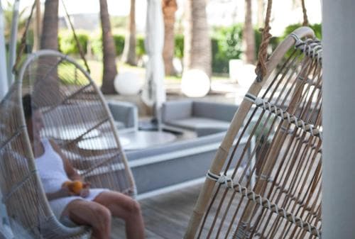 A serene tropical resort scene features a person relaxing in a woven hammock chair near a modern poolside area, surrounded by lush palm trees, ideal for leisure travelers seeking tranquility.