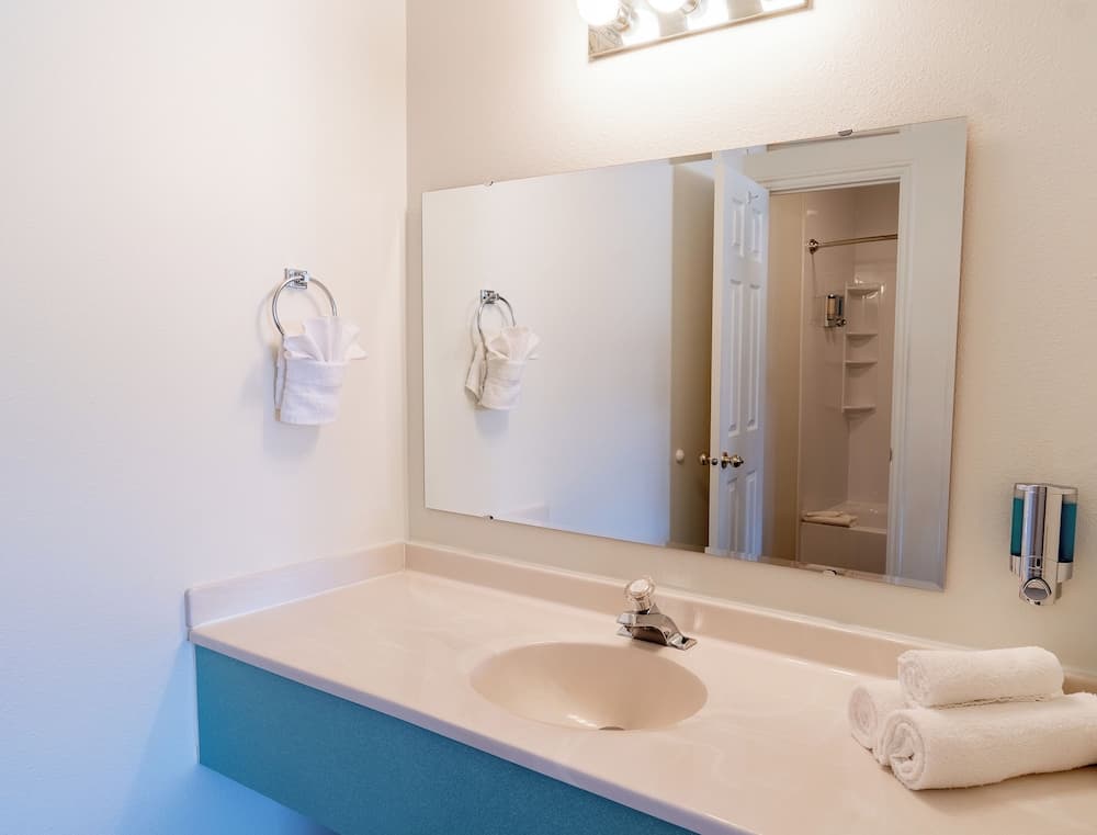 A clean and inviting hotel bathroom features a white sink countertop with neatly rolled towels, a large mirror reflecting a shower area, and a towel ring holding a fresh hand towel, ideal for travelers seeking comfort and convenience.