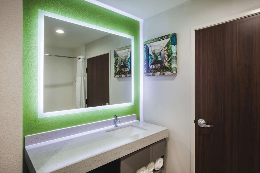 A modern hotel bathroom features a sleek white vanity with a green-lit mirror, tropical artwork on the walls, and neatly folded towels, creating a refreshing and inviting atmosphere ideal for travelers seeking comfort and style.