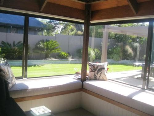 A cozy corner window seat with cushions overlooks a serene garden with lush greenery, ideal for a tranquil retreat or a relaxing reading spot during a vacation.