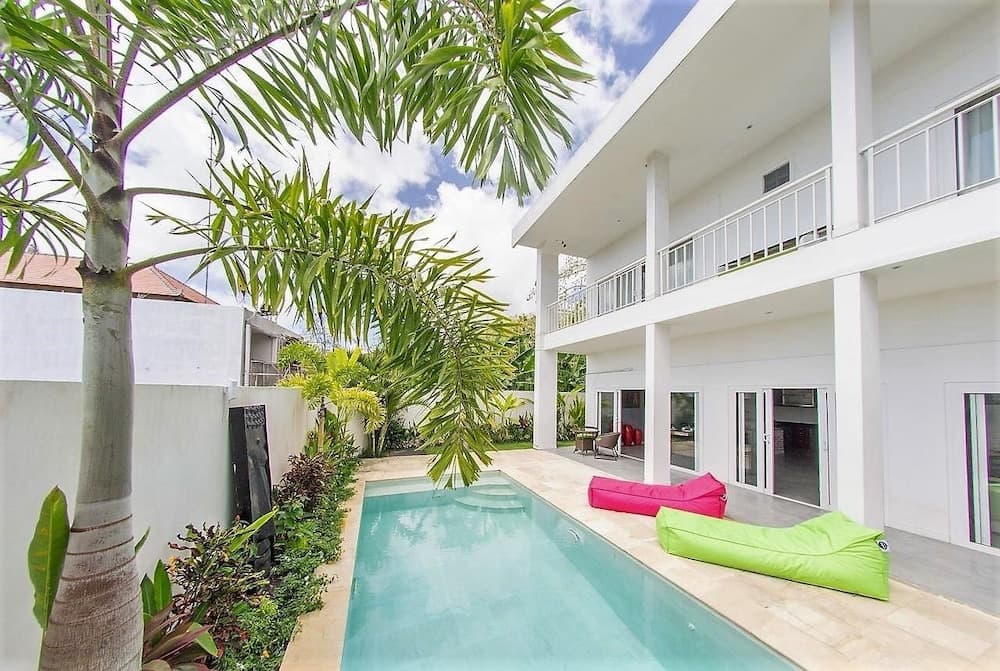 A serene tropical villa features a tranquil poolside area with colorful bean bags, lush palm trees, and modern architecture, ideal for relaxing vacations or luxurious getaways.
