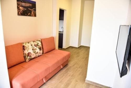 A cozy apartment interior features an orange sofa with floral cushions, wooden flooring, and a mounted TV, ideal for travelers seeking comfort in a modern living space.