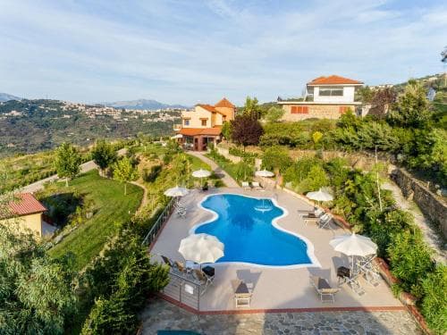 A serene hillside villa features a uniquely shaped pool surrounded by lush greenery, white umbrellas, and cozy seating, ideal for a tranquil vacation retreat.