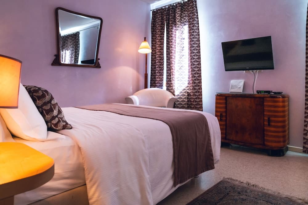 A cozy hotel room with soft purple walls, a neatly made bed adorned with patterned pillows, a vintage wooden cabinet under a mounted TV, and warm lighting creates an inviting atmosphere ideal for travelers seeking comfort and relaxation.