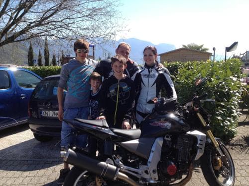 A family poses happily beside a sleek motorcycle in a sunny parking lot near lush greenery and distant mountains, ideal for adventure travelers seeking scenic routes and family-friendly destinations.
