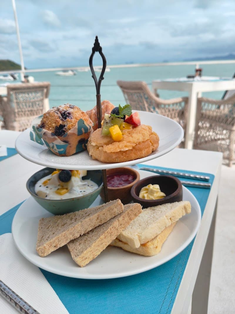 A serene beachside dining experience at Tembo Beach Club & Resort features a delightful tiered plate of pastries, fresh fruit, and breads set against the tranquil backdrop of Bangrak Beach in Thailand, perfect for travelers seeking a luxurious seaside retreat.
