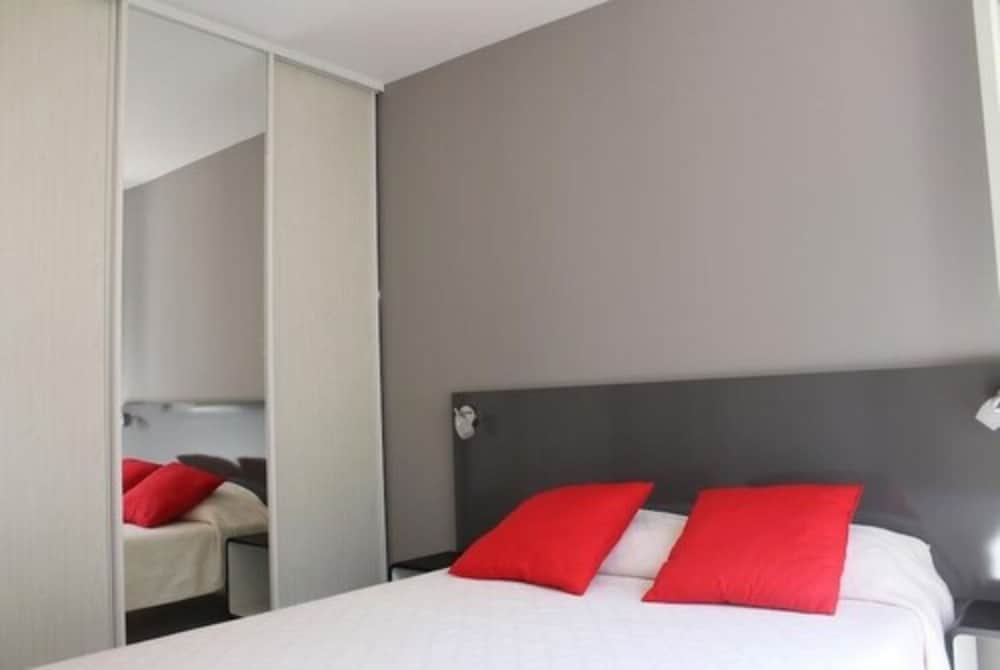 A cozy hotel bedroom features a modern bed with vibrant red pillows against a sleek gray headboard, mirrored closet doors reflecting the room's comfort, ideal for travelers seeking a stylish yet minimalist retreat.