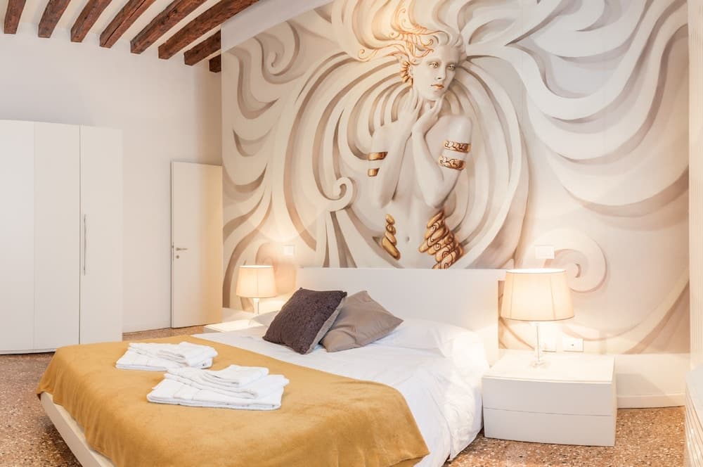 A serene bedroom features an artistic mural of a woman with flowing hair as the focal point, complemented by modern furnishings, warm lighting, and exposed wooden beams, ideal for travelers seeking a cozy and stylish accommodation.