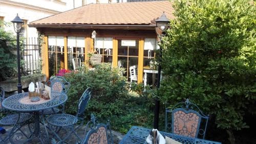A cozy outdoor café scene features ornate metal chairs and tables set amidst lush greenery, evoking a serene and inviting atmosphere perfect for leisurely dining or relaxing getaways.