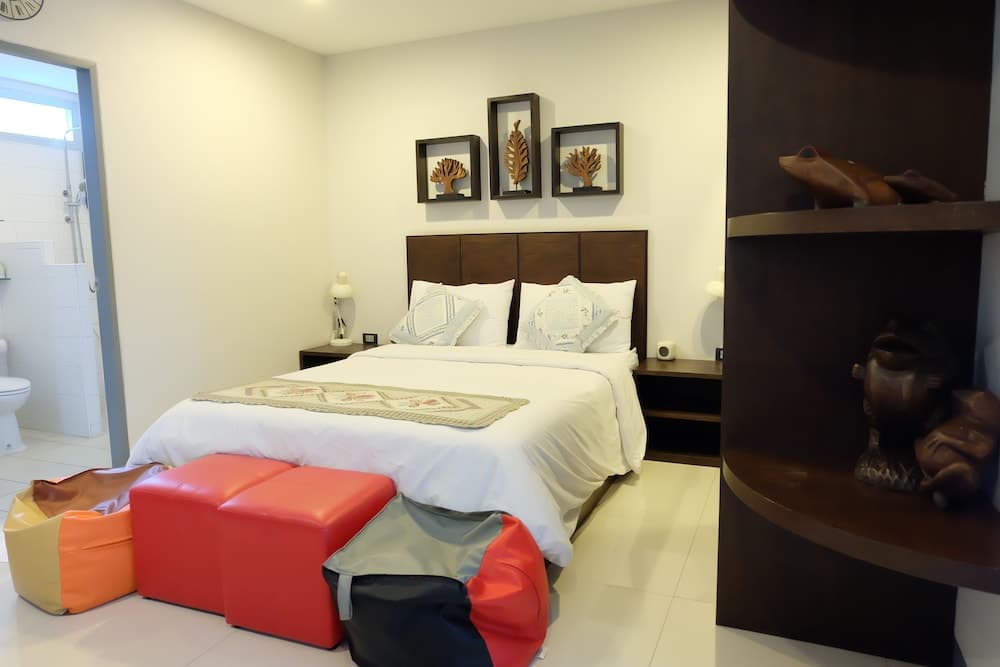 A cozy hotel bedroom features a neatly made bed adorned with decorative pillows, vibrant red ottomans, wooden shelves displaying unique sculptures, and framed wall art, creating a welcoming atmosphere ideal for travelers seeking comfort and style.